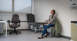 Full length wide view of a mid adult woman sitting in a doctors examination room waiting to be seen, she is holding her arm and looking up, feeling worried. She is wearing casual clothing at the hospital in Newcastle, England.