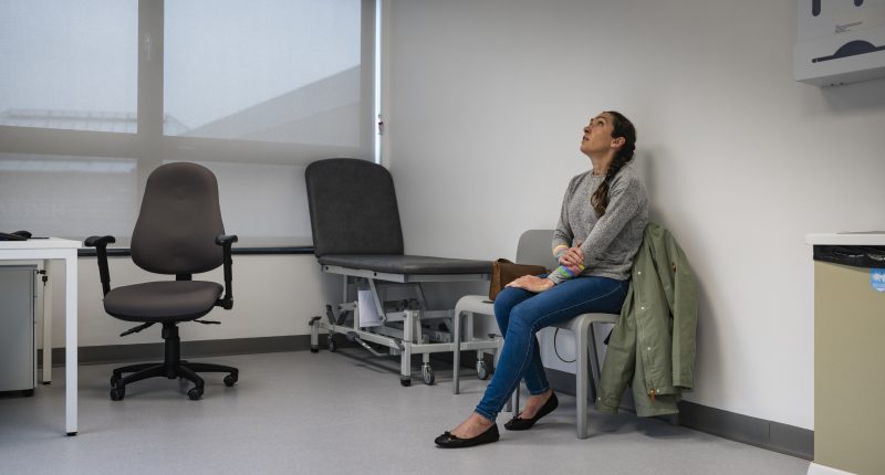 Full length wide view of a mid adult woman sitting in a doctors examination room waiting to be seen, she is holding her arm and looking up, feeling worried. She is wearing casual clothing at the hospital in Newcastle, England.